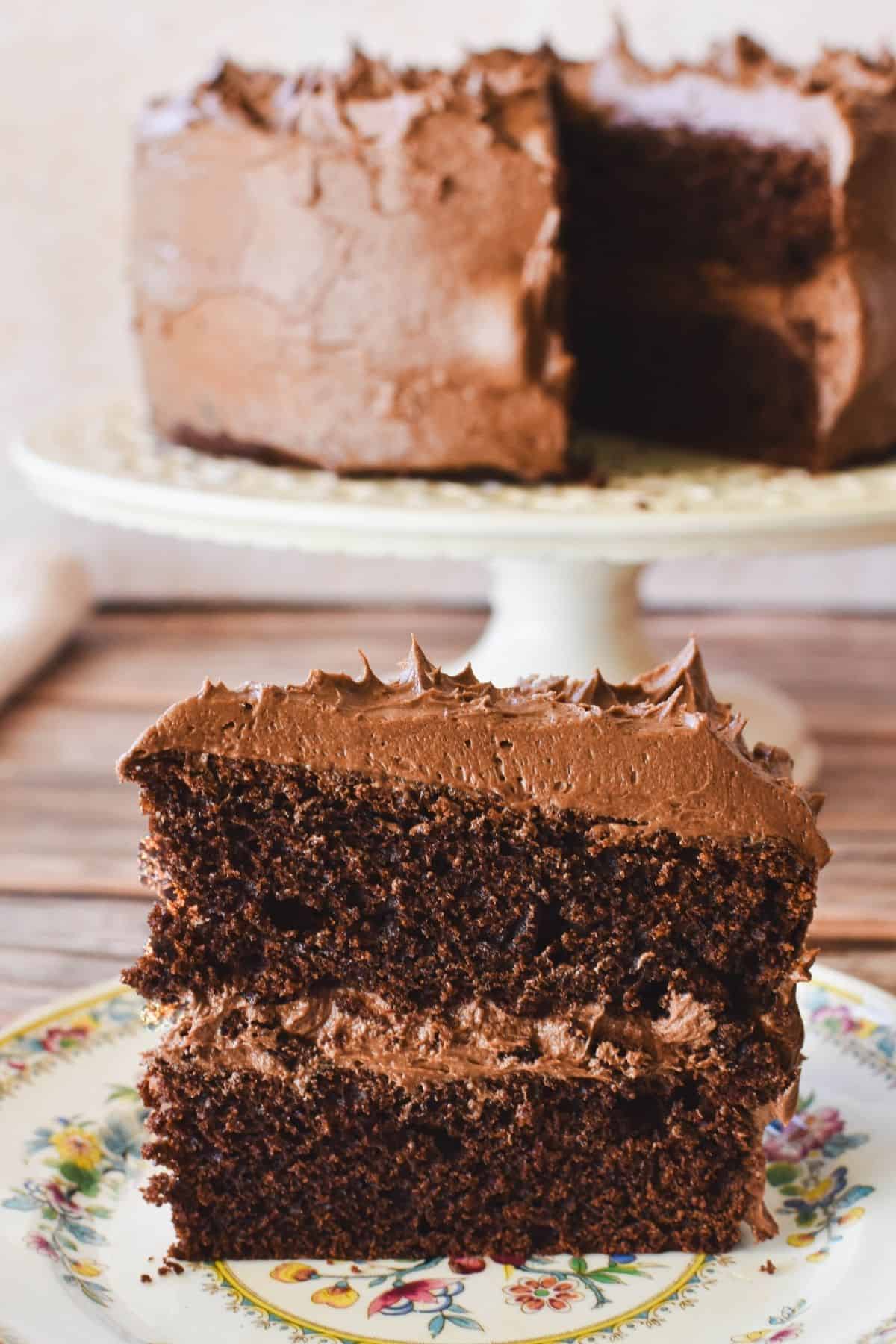 Slice of eggless chocolate cake on plate with whole cake in background.